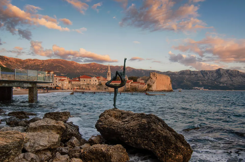 Ballerina statue on rocks with Budva's old town walls glowing at sunset behind it