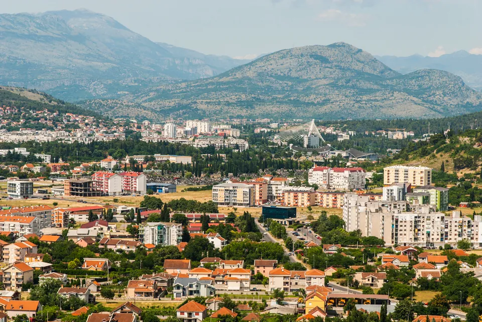 Podgorica's residential rooftops spread beneath green hills and layered mountains in haze