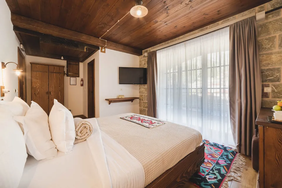 Bedroom with stone wall, white linen, wooden ceiling, tall window and woven rug