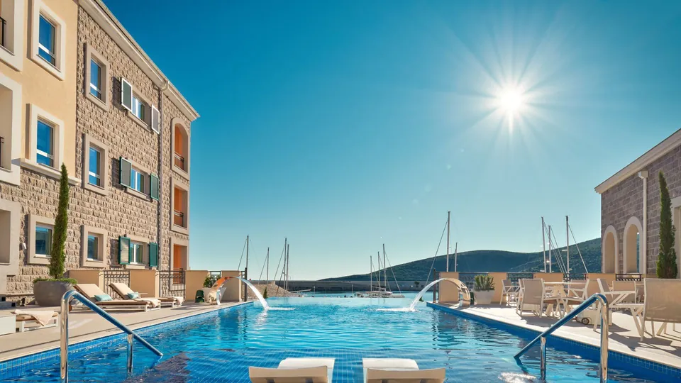 Hotel pool with loungers, water jets, marina masts, hills and bright sun