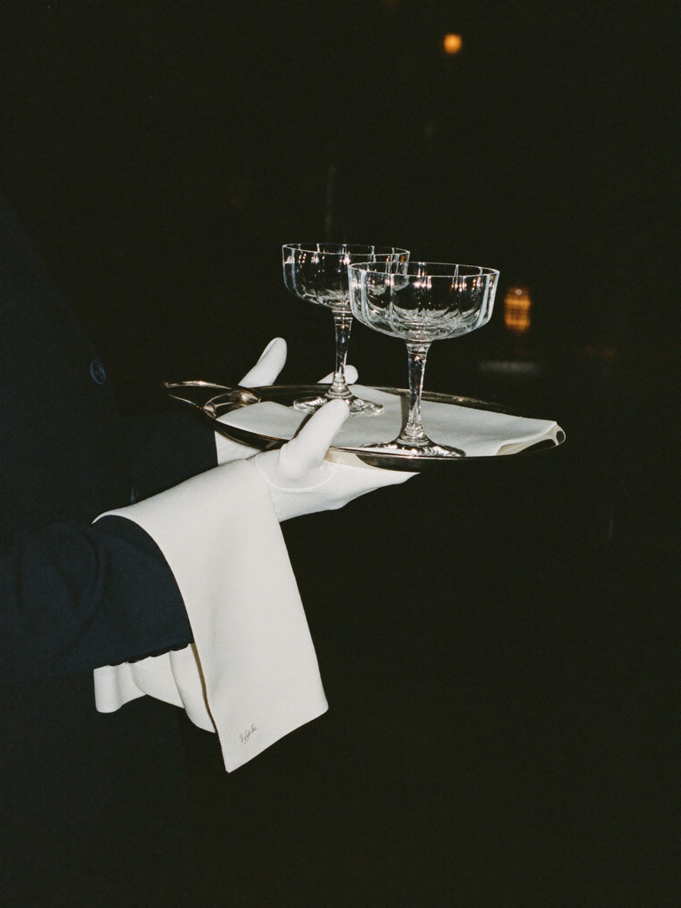A gloved hand carrying a silver tray with two coupe glasses
